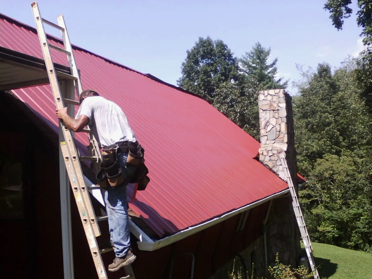 Roofer on a ladder beside a red metal roof and stone chimney — Soffit & Fascia Repair by Roofing Brothers in Morrisdale